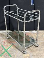 Front side view of the chrome bamboo bar cart showing the two smoked glass shelves and caster wheels on a concrete floor.