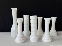 Photo showing all seven white milk glass bud vases together in a group against a black background, highlighting their assorted shapes and heights.
