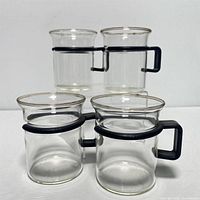 Top-down front view of four clear glass coffee cups arranged in a diamond shape with black rectangular handles and black ring accents embroidered around upper midsection.