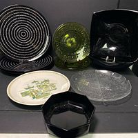Photo showing the entire set of 10 plates and dishes against a black background, displaying variety in style and material including black, green, and clear glass dishes and one decorative ceramic plate.