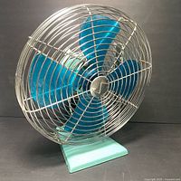 Front view of the Superior Electric vintage fan showing blue metal blades inside silver wire cage and turquoise base.