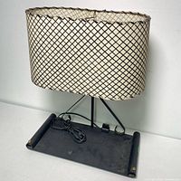 Full view of lamp base and shade showing black metal base with curled decorative arms and the oval white fiberglass shade with black crosshatched design.