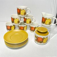 Photo showing 6 tea cups stacked in a pyramid shape behind 6 mustard yellow saucers, creamer pitcher and sugar jar with lid, all decorated with a vintage fruit motif.