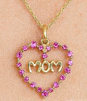 Gold heart-shaped pendant with pink stones outlining the heart and 'Mom' text inside, shown on textured pink background.