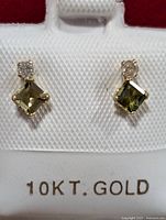Front view of the pair of 10K gold earrings with green tourmaline stones and diamond accents on white textured card marked 10KT GOLD.