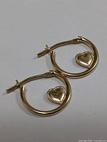 Pair of small 14 karat gold hoop earrings with heart-shaped pendants on a plain background.