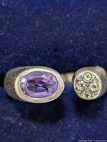 Close-up image showing the oval faceted amethyst and smaller green gemstone cluster on silver ring against a dark blue background.