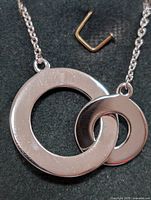 Clear close-up photo of the sterling silver necklace pendant showing two interlocking circles and part of the chain, taken on a dark background with a small metal U-shaped object nearby.