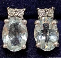 Close-up front view of two silver earrings with oval blue topaz stones and small clear accent stones, placed on dark background.