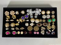 Tray holding over 20 pairs of assorted vintage costume earrings in gold-tone and silver-tone metals, featuring faux pearls, rhinestones, and colorful accents. Some earrings show missing stones, minor wear visible.
