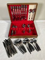 Two sets of flatware arranged inside and in front of the wooden chest showing ornate handles and multiple utensils including knives, forks, tablespoons, and teaspoons.