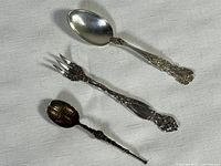 Three sterling silver cutlery pieces including a spoon, a fork, and a small ornate spoon on white textured fabric surface
