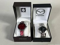 Two ladies wristwatches displayed side by side in original boxes: Anne Klein with red face and pink band; Mazda with black face and silver metal band.