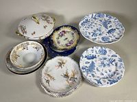 Top-down view of assorted vintage china including a covered vegetable bowl, various decorated plates including blue and white floral, and multi-colored floral patterns.