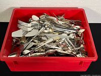 Plastic red bin filled with assorted silver-toned vintage cutlery and utensils including forks, knives, spoons, tongs, ladles, and cake cutter.