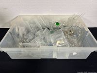Clear plastic bin containing various loose glass items including stemware, a creamer, and small plates.