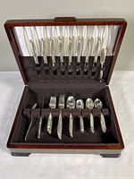 Open wooden chest displaying ten dinner knives held upright in loops on satin interior behind the lid, and various forks and spoons arranged on felt base inside the box.