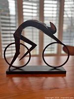 Front angled view of the bronzed abstract bicyclist statue showing the rider's form hunching over the frameless bicycle with two large circular wheels and a black rectangular base.