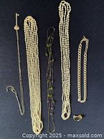 Photo of five necklaces and two rings laid out on a dark textured surface. Includes multi-strand faux pearl necklaces, a beaded necklace with green accents, a chain-link style necklace, and two decorative rings.
