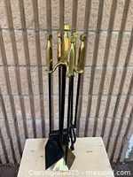 Full view of the fireplace tool set with six tools standing upright on the stand against a beige brick wall and white baseboard background.