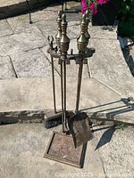 Full view of antique brass fireplace tools on stand showing poker, brush, shovel, and square base stand on a paved floor.