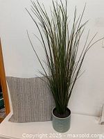 Artificial grass plant with tall slender green stems in light blue ceramic pot with circular pattern and decorative beige woven pillow behind.
