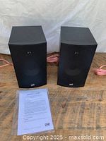 Front view of both PSB Alpha B1 black bookshelf speakers sitting side by side with owner's guide in front on wooden surface with white background.
