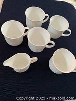 Top-down view of 5 white ceramic coffee cups and 1 creamer on dark textile surface, showing ribbed pattern and cup handles.