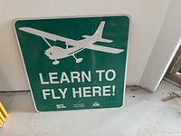 Green square Learn to Fly Here metal sign leaning against wall