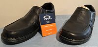 Pair of black men's slip-on loafers angled front view showing moc toe stitching and padded collar with original hang tag attached.