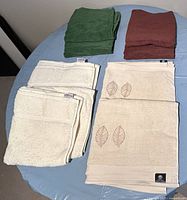 Four sets of folded towels on a round surface: green, brown, beige with embroidered leaves, and ivory.