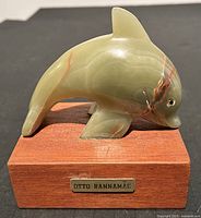Front view of the dolphin sculpture showing the smooth jade-like stone with natural color streaks and wooden base with metal artist plaque.