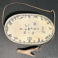 Top view of oval-shaped hand-crafted Inuit drum featuring black indigenous artwork on drum skin and attached wooden drumstick on leather lanyard.