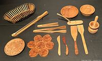 Full view of entire lot with bread basket, bread cutter, wooden bowls, juniper mortar and pestle, trivet, plate, tongs, spatulas, and spreaders arrayed on black surface