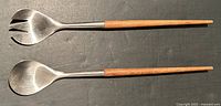 Pair of MCM teak handled stainless steel salad servers laid side by side, showing one fork-style and one spoon-style server, both approximately 13 inches long.
