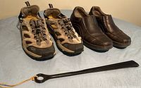 Two pairs of men's shoes side by side with black shoe horn in front on a blue surface. Brown Clarks loafers and tan/black Sportsman hikers/runners with open sides.