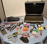 Overview of lot showing metal briefcase, black shredder, staplers, scissors, tape rolls, and assorted office supplies on table