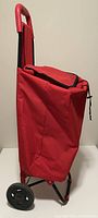 Red grocery pull buggy standing upright showing side view, black wheels, fabric bag with flap closed.