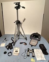 Lot arranged with Top-Man tripod extended vertically. Canon PowerShot SX10IS camera with lens cap, mini tripod, Bushmaster binoculars with case, carrying case, wrist strap, and instruction manuals all spread out on flat surface.