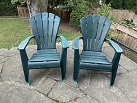 Two green resin Muskoka chairs outside on a stone patio, showing front view, both chairs with slatted backs and broad armrests. One chair shows a visible crack repaired on the hand-rest.