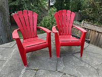Two red resin Muskoka (Adirondack) chairs positioned outdoors on a stone patio with greenery background. Both chairs shown side by side from front angle.