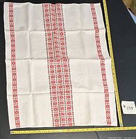 Red-striped linen-like fabric tablecloth laid flat showing central and edge pattern with measuring tape along edges for size reference.