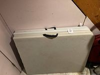 Two white folding tables folded flat, stacked against a corner wall, showing the top surface and black handles.