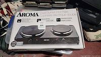 Photo showing the Aroma AHP-312 dual burner hot plate in its original box from a front angle.