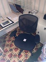 Black office chair from a top angle showing mesh backrest and seat, armrests, part of surrounding area including desk and floor.