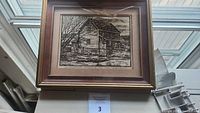 Framed woodcut print of an old grist mill by Carl Percy Martin displayed on a surface next to a window.