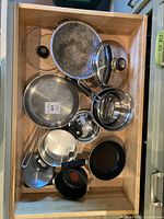 Top view of cookware pieces including stainless steel pots, pans, strainer pan, and lids inside a wooden drawer