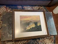 Full view of the signed framed landscape painting showing the frame and the artwork inside placed on a carpet.
