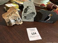 Three soapstone carvings displayed on a table showing the variety of painted scenes including horses, a baker, and a puffin bird