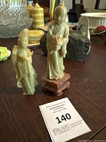 Two jade sculptures placed on a table with various other items in the background. One statue is mounted on a wooden base, the other lacks its base.